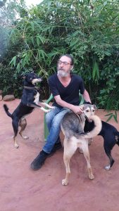Image of man with glasses and a white beard, sitting in a garden and playing with three dogs