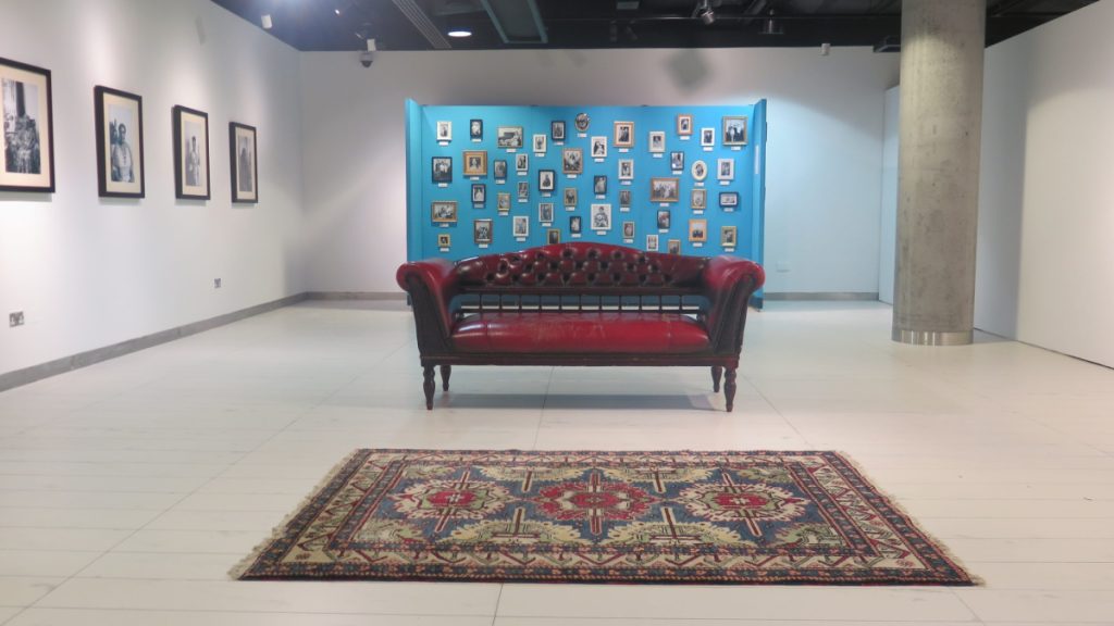Image of a white gallery space. There are black and white photos on the left wall. In front, there is an antique red leather sofa, behind that is a blue wall with lots of smaller framed black and white photographs, displayed salon style