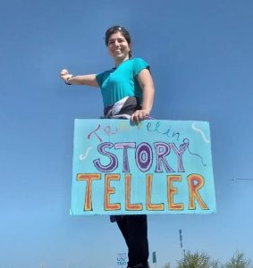 Image of a woman hitch-hiking, she is standing against a blue sky and has a sign saying 'traveling storyteller' held in front of her