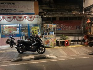 A dark street, there are some parked mopeds by the side of the road. There is a small food stall next to one of the mopeds.