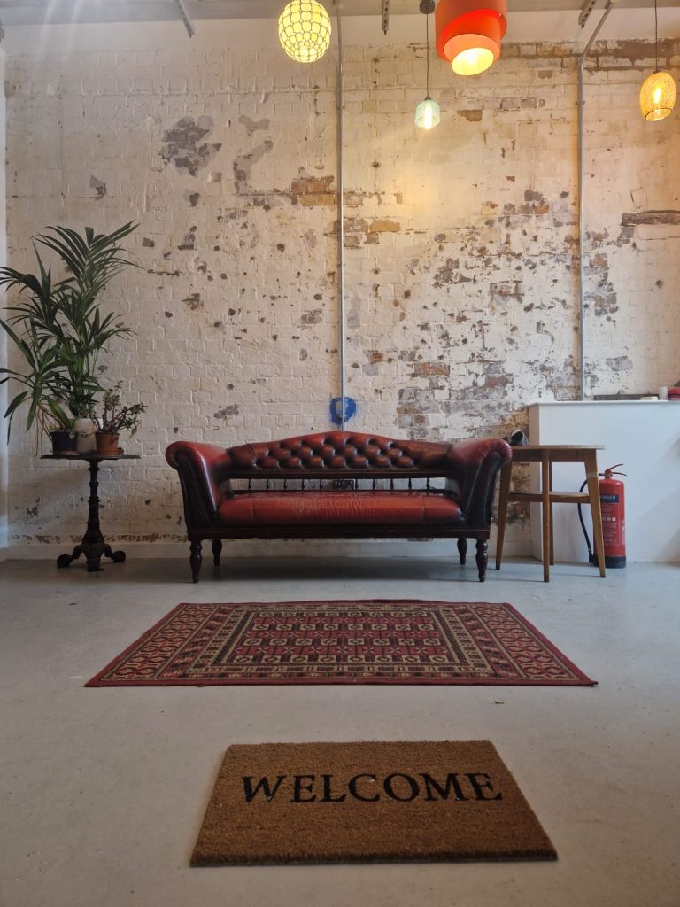 An ornate red sofa, with a large pot plant to one side and an oriental rug in front. It is against a wall with distressed white and grey paint.