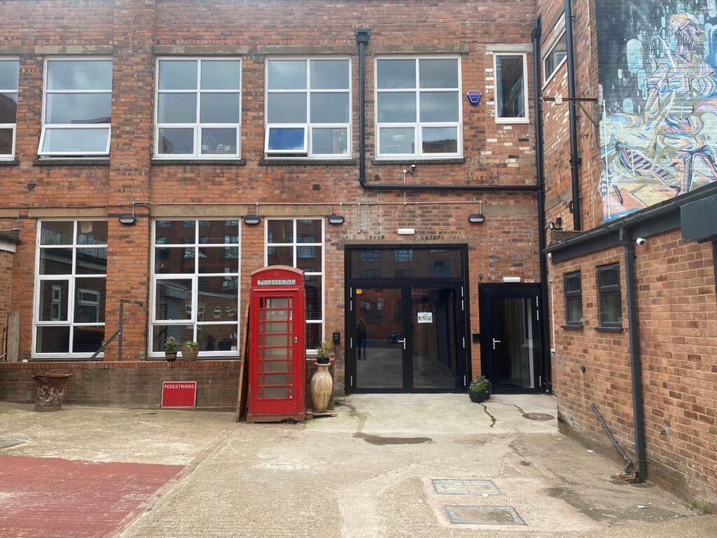 A picture of a twwo storey, industrial style building. There are many windows, and glass fronted doors. An old red telephone box stands in front of it.