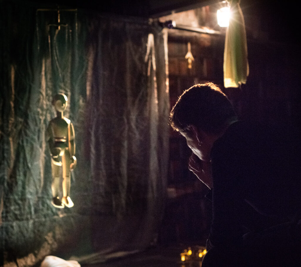 A picture of a dark theatre space, with Freda bent down in front of a wooden puppet