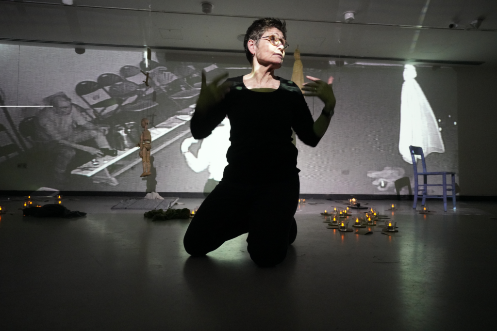 A picture of a woman standing, posed in a theatre space, behind her is a large, black and white projection. There are many tiny lights on the floor, and a wooden puppet is hanging in front of the projection screen