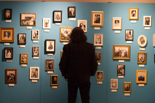 Image of a man from the back, he has long dark hair. He is standing in front of a blue wall on which there are many small black and white framed photographs.
