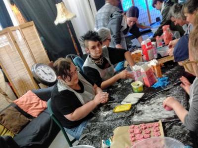 Image of around twenty people sitting around a long table making sweets. The table is messy and has lots of ingredients and plastic bowls scattered along it