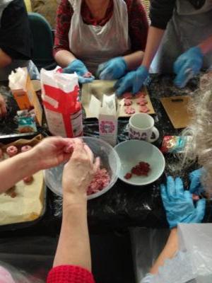Image of five people's hands encased in plastic gloves, all busy making cookies on a table covered in ingredients and plastic bowls.